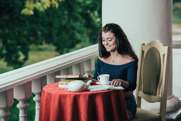 libro de lectura de la chica joven mientras que bebe el café en el día soleado que se sienta en el balcón.