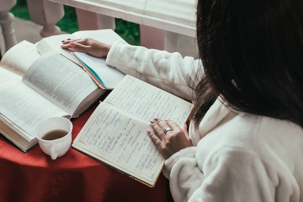 libro de lectura de la chica joven mientras que bebe el café en el día soleado que se sienta en el balcón.