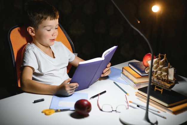 Libro de lectura de colegial concentrado en la mesa con libros, planta, lámpara, lápices de colores, manzana y libro de texto