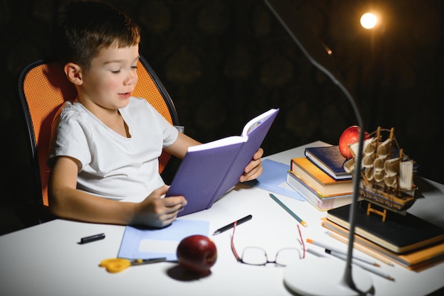 Foto libro de lectura de colegial concentrado en la mesa con libros, planta, lámpara, lápices de colores, manzana y libro de texto