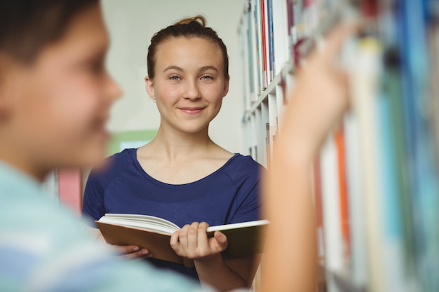 Libro de lectura de colegiala sonriente en la biblioteca de la escuela