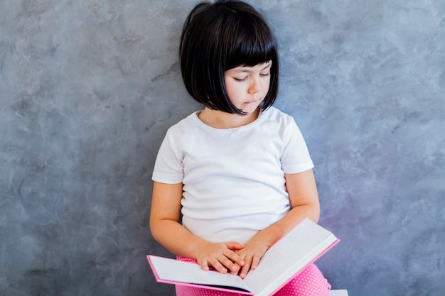 Foto libro de lectura lindo de la niña del pelo negro por la pared