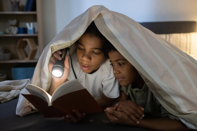 Libro de lectura de niña afroamericana al hermano menor bajo la luz de la linterna