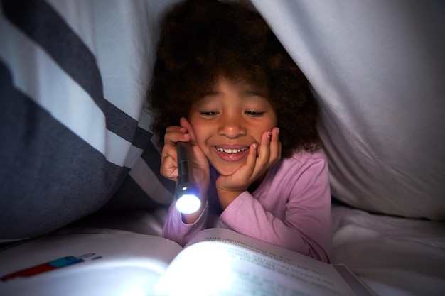 Libro de lectura de niña bajo el edredón en la cama por la noche con antorcha