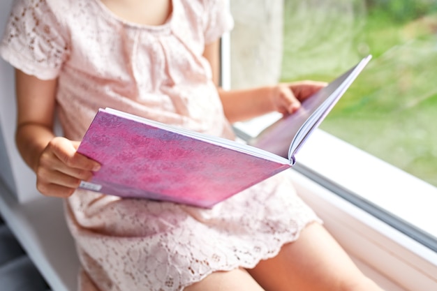 Libro de lectura de niña linda en casa, en el alféizar de la ventana, niño estudiando en casa, cuarentena, quedarse en casa, estilo de vida.