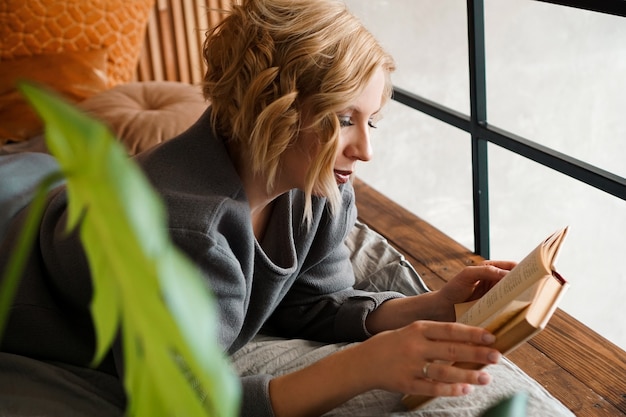 Libro de lectura de niña rubia en la cama - acogedora habitación con plantas verdes