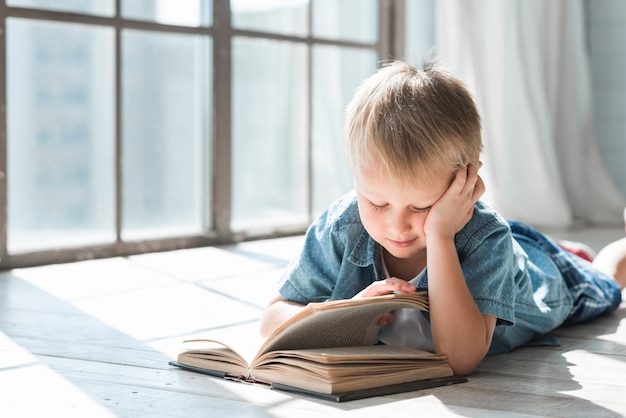 Libro de lectura rubio del muchacho cerca de la ventana en la luz del sol