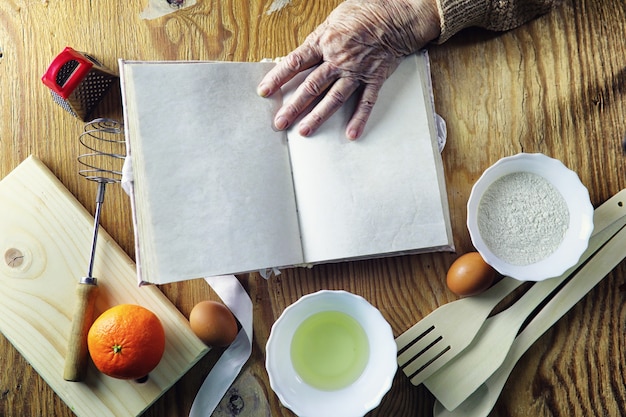 Libro de recetas abierto en manos de una anciana frente a una mesa con utensilios