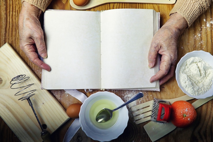 Foto libro de recetas abierto en manos de una mujer mayor frente a una mesa con utensilios