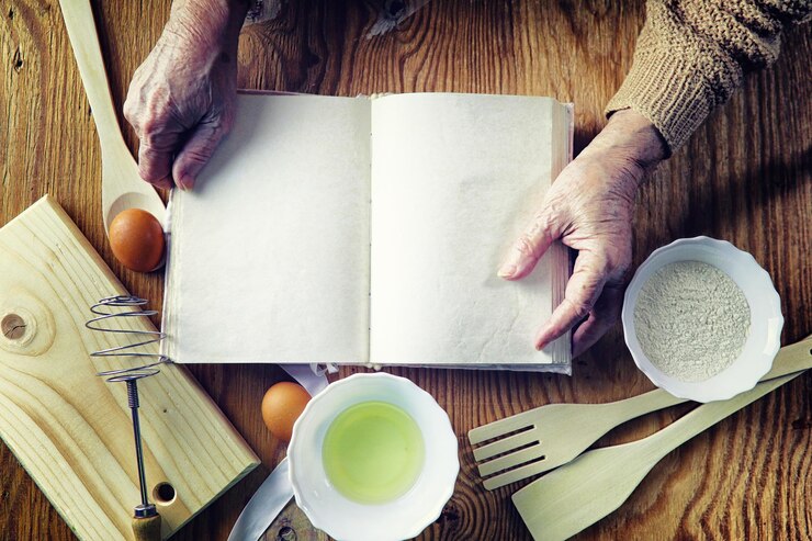 Foto libro de recetas abierto en manos de una mujer mayor frente a una mesa con utensilios