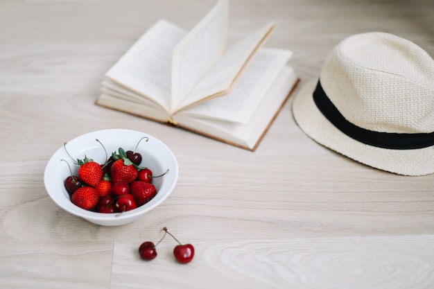 Libro de sombrero de traje de baño y cerezas y fresas frescas sobre superficie de madera