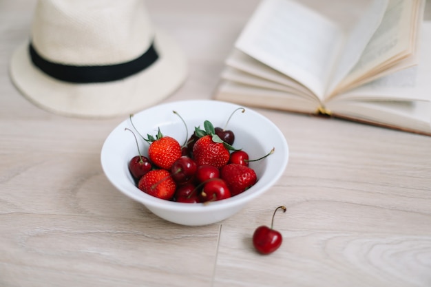 Libro de sombrero de traje de baño y cerezas y fresas frescas sobre superficie de madera