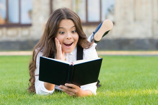 Libro con sorpresa inesperada Niño sorprendido lee un libro sobre hierba verde Niña leyendo con emoción sorpresa en la cara Linda colegiala pequeña mantiene la boca abierta de gran sorpresa Concepto sorpresa