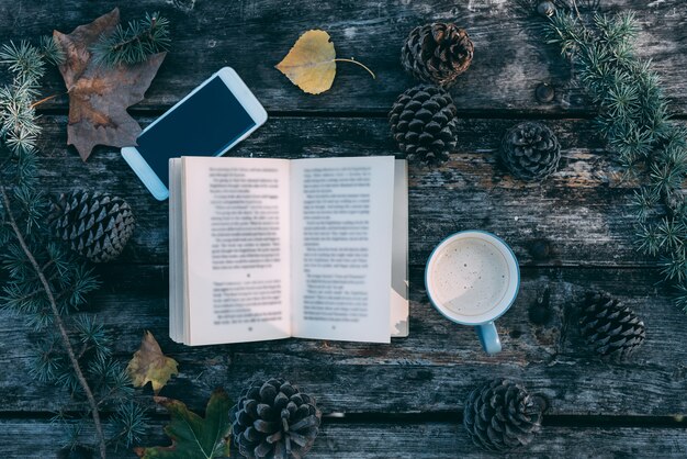 Libro y teléfono móvil en una mesa de madera con café y pinos al aire libre