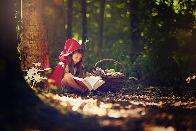 Los libros ayudan a expandir el mundo y la imaginación de los niños Foto de una niña con una capa roja leyendo un libro en el bosque