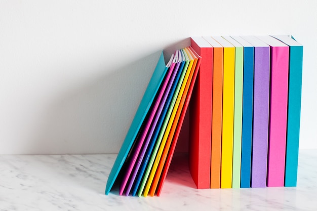 Los libros coloridos se describen con los colores del arco iris. Apilados de los libros en un estante cerca de la pared blanca.