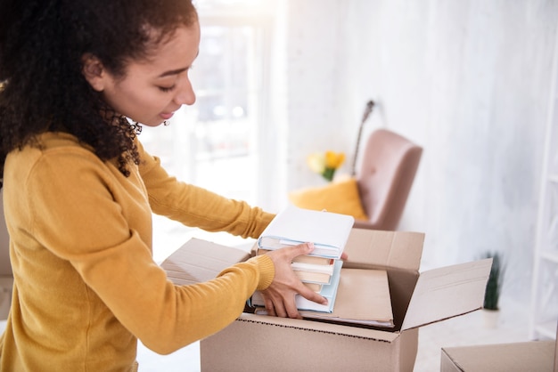 Libros necesarios. Encantadora niña de cabello rizado que coloca una pila de libros en la caja mientras se prepara para mudarse de un piso viejo