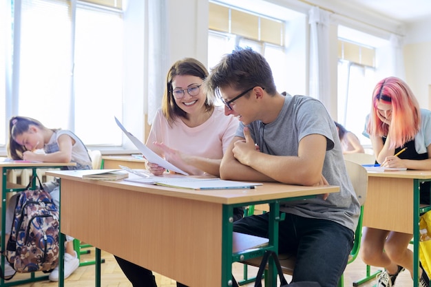 Lição na classe de estudantes do ensino médio, professora sentada na mesa com adolescente estudante do sexo masculino.