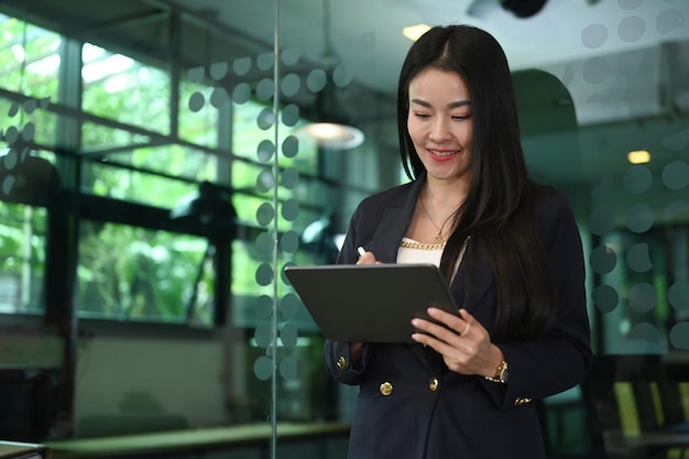 Líder de equipe feminina sorridente em pé no escritório corporativo e usando tablet digital