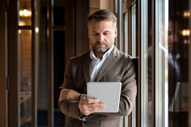 Líder empresarial serio en ropa formal que se concentra en la red mientras mira la pantalla táctil mientras está de pie junto a la ventana