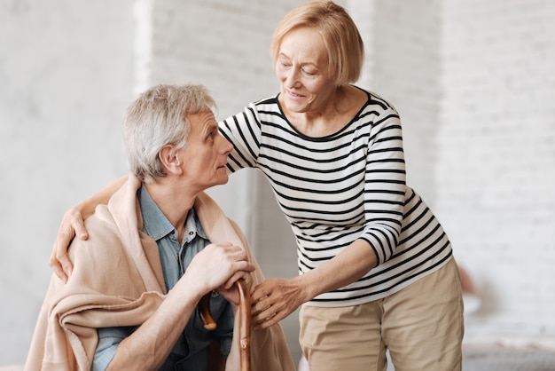 Liebe heilt alles. Nette süße ältere Frau, die dafür sorgt, dass sich ihr Mann wohl und warm fühlt, während sie ihm eine Decke zur Verfügung stellt und ihn ein wenig umarmt