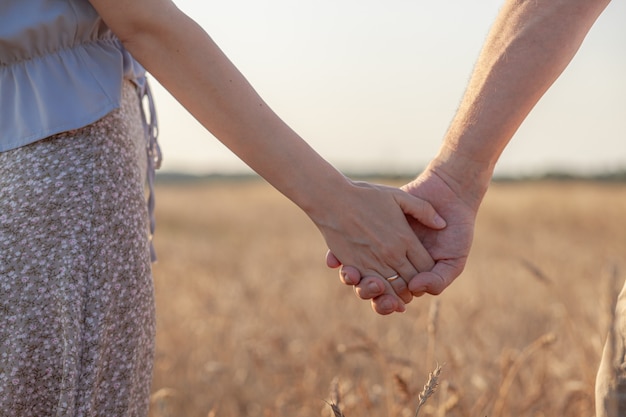 Liebe Konzept. Ein Paar, das während des Sonnenuntergangs die Hand hält, ein Symbol für Liebe und glückliche Beziehung. Ein junges verliebtes Paar geht bei Sonnenuntergang durch ein Weizenfeld, hält Händchen und schaut auf den Sonnenuntergang