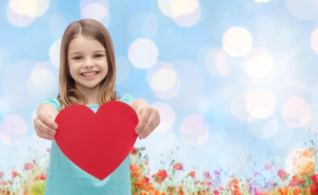 Liebe, Nächstenliebe, Feiertage, Kinder und Personenkonzept - lächelndes kleines Mädchen mit rotem Herzen über blauem Licht und Mohnfeldhintergrund