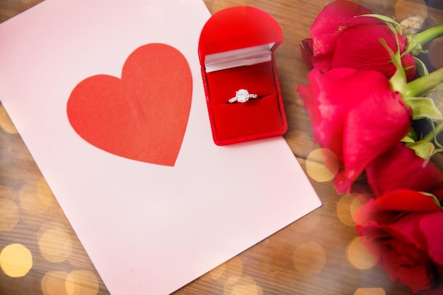 Liebe, Vorschlag, Valentinstag und Feiertagskonzept - Nahaufnahme der Geschenkbox mit Diamant-Verlobungsring, roten Rosen und Grußkarte auf Holz