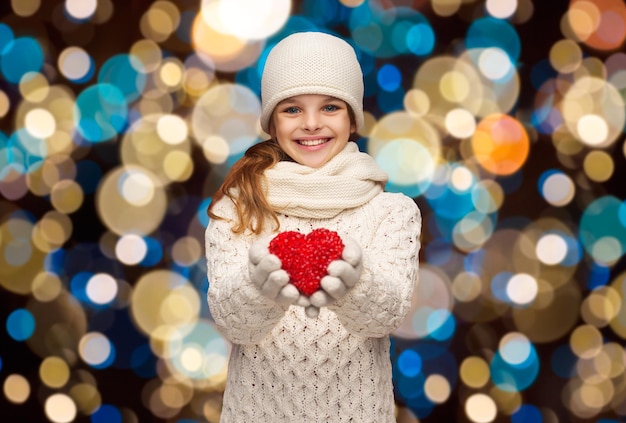 Liebe, Wohltätigkeit, Weihnachten, Feiertage und Menschen Konzept - glückliches Mädchen in Winterkleidung mit rotem Herzen über Lichtern Hintergrund