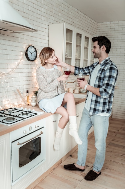 Liebendes Paar. Attraktiver junger dunkelhaariger Mann, der ein kariertes Hemd trägt und seine Frau, die sich romantisch fühlt, während sie Wein trinkt