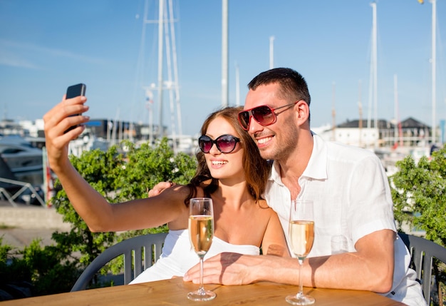 Liebes-, Dating-, Menschen- und Urlaubskonzept - lächelndes Paar mit Sonnenbrille, Champagner trinken und Selfie im Café machen