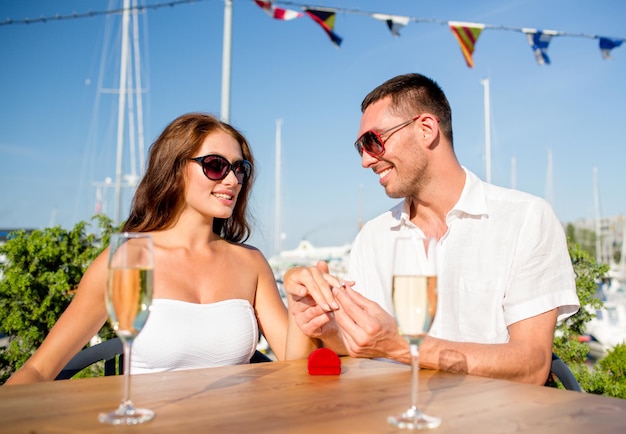 Liebes-, Dating-, Menschen- und Urlaubskonzept - lächelndes Paar mit Sonnenbrille mit Champagner und kleiner roter Geschenkbox, die im Café den Ehering auf den Finger legt