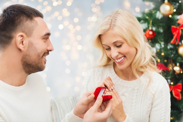 Liebes-, Paar-, Heiratsantrags-, Feiertags- und Menschenkonzept - glücklicher Mann, der der Frau einen Diamant-Verlobungsring in einer kleinen roten Schachtel über Weihnachtsbaum und Lichterhintergrund schenkt
