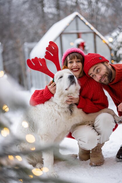 Liebes Paar umarmt mit ihrem niedlichen Hund trägt Spielzeug Hirsch Hörner im schneebedeckten Hinterhof Junge Familie verbringt glückliche Winterzeit zusammen im Freien
