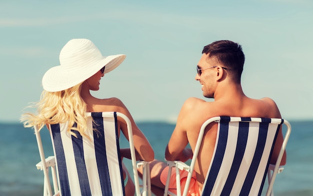 Liebes-, Reise-, Tourismus-, Sommer- und Menschenkonzept - lächelndes Paar im Urlaub in Badebekleidung, das auf Stühlen sitzt und sich von hinten am Strand sonnt