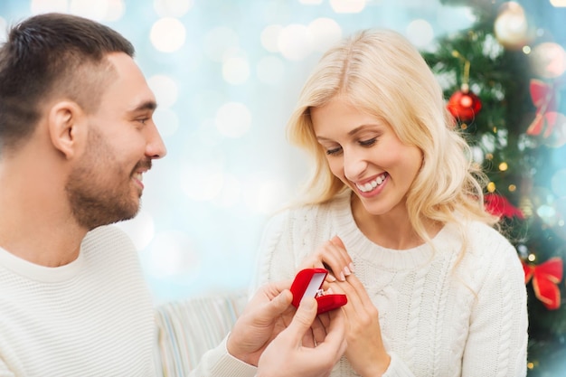Liebes-, Weihnachts-, Paar-, Vorschlags- und Personenkonzept - glücklicher Mann, der der Frau über blauen Feiertagen einen Diamant-Verlobungsring in einer kleinen roten Schachtel gibt, beleuchtet den Hintergrund