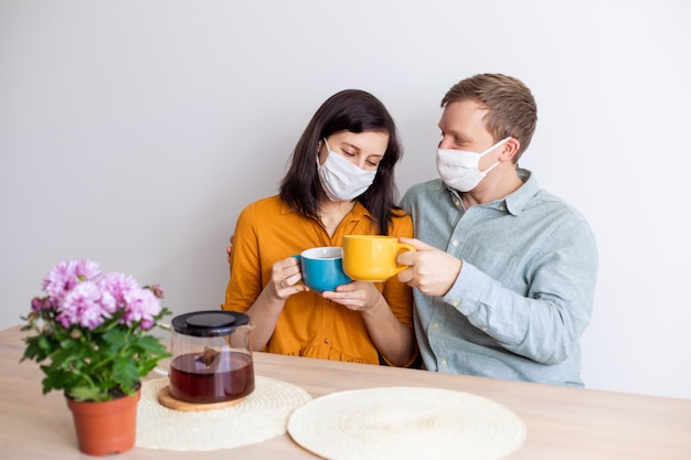 Liebesgeschichte Familie Ehemann Ehefrau Freund Freundin in sterilen Masken unter Quarantäne gestellt. Normales Leben Trinken Sie Tee mit Coronavirus. Lebensstil COVID-19. Quarantäne-Virenschutz Sterilität nach Hause zusammen