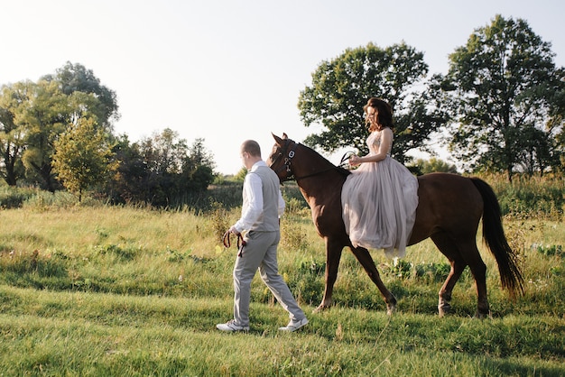 Liebesgeschichte und Hochzeit nahe dem Fluss in einem Feld bei Sonnenuntergang mit braunem Pferd. Die Braut im luftigen Kleid hat die Farbe einer staubigen Rose. Beiges Kleid mit Glitzern. Braut und Bräutigam umarmen und küssen.