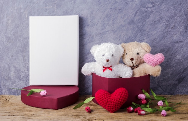 Liebeskonzept des Teddybären in der roten Herzgeschenkbox auf hölzerner Tabelle und leerem Segeltuchrahmen