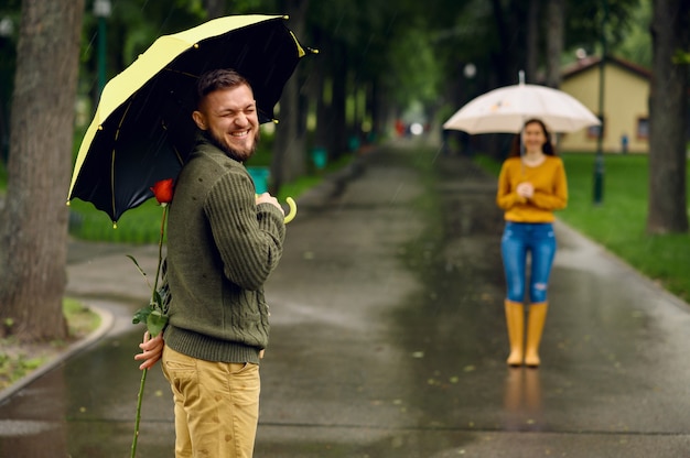 Liebespaar mit Regenschirmen trifft sich im Park, regnerischer Tag. Mann mit roter Rose wartet auf seine Frau auf Gehweg, nasses Wetter in Gasse