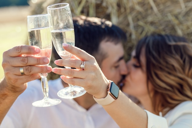 Liebespaar trinkt Weinrebe bei Sonnenuntergang auf einem Weizenfeld in der Nähe von Heuhaufen. Ein Freund und ein Mädchen bei einem romantischen Date, sitzen in der Nähe eines Heuhaufens und trinken Champagner aus Gläsern.