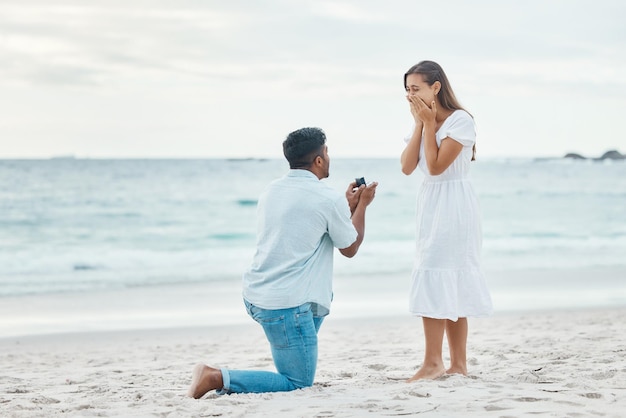 Liebespaar und Strand-Verlobungsvorschlag für Ehepartnerschaft und Engagement auf Meeressand Wow schockierte und überraschte Frau mit romantischem Mann auf einem Knie, der einen gewerkschaftlichen Ehering präsentiert