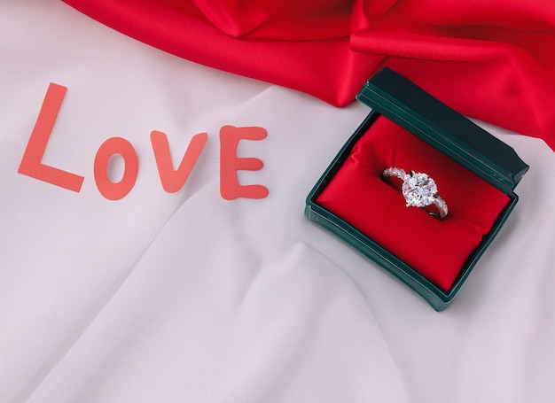 Liebeswort auf weißem Stoffhintergrund mit Verlobungsdiamantenring in einer Ringbox mit Rose