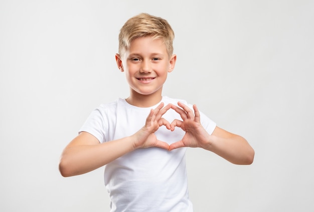 Liebeszeichen. Zuneigung Mitgefühl. Freundlicher blonder Junge, der Herzgeste einzeln auf neutraler Wand zeigt.