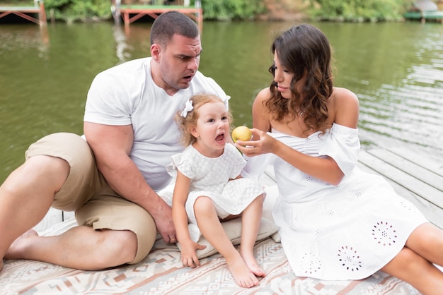 Liebevolle Familie sitzt auf einer Brücke am Fluss Mama Papa und kleine Tochter hatten ein Picknick am Wasser Mama füttert ihre Tochter mit einem Apfel die Tochter macht ein Gesicht