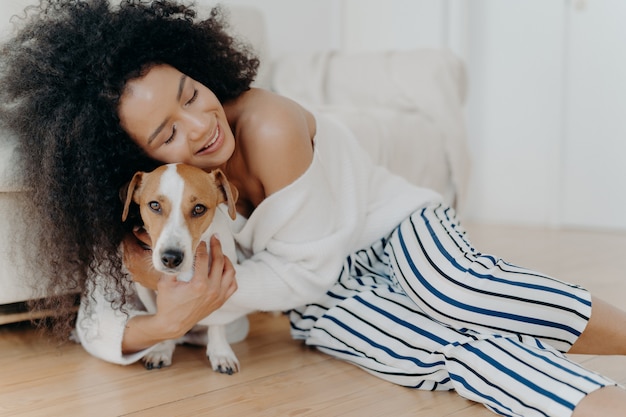 Liebevolle junge Frau umarmt Hund mit Liebe und Sorgfalt, hält die Augen vor Vergnügen geschlossen, lächelt sanft