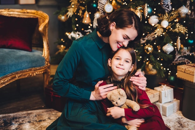 Liebevolle Mutter umarmt ihre kleine Tochter, während sie in der Nähe des geschmückten Weihnachtsbaums zusammensitzen Kleines entzückendes Mädchen, das froh ist, Neujahrsgeschenk von ihrer Mutter zu erhalten Beziehungskonzept