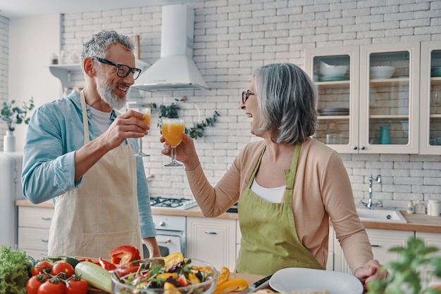 Liebevolle Seniorenpaare in Schürzen, die sich gegenseitig mit Orangensaft anstoßen und ein gesundes Abendessen zubereiten, während sie Zeit zu Hause verbringen