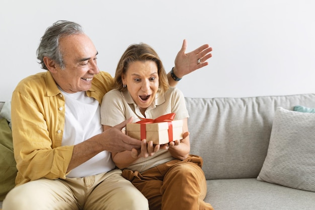 Liebevoller älterer Ehemann, der der erstaunten Ehefrau ein Überraschungsgeschenk macht, eine glückliche alte Frau wird unerwartet