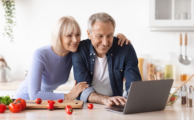 Liebevolles älteres Paar, das zu Hause mit Laptop zusammen kocht